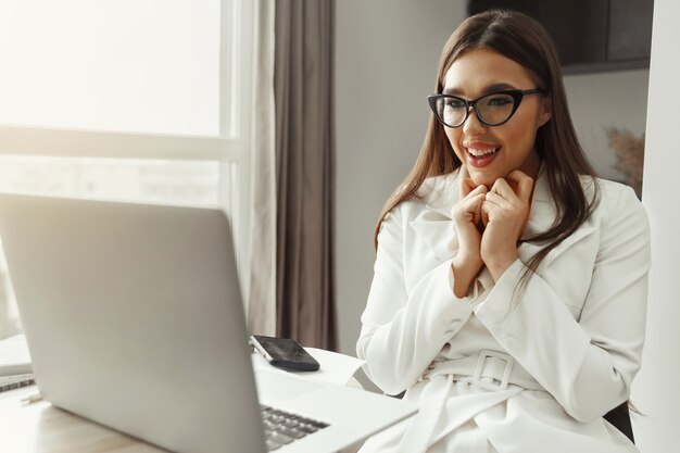 Piękna Szczęśliwa Młoda Bizneswoman, Kobieta Za Pomocą Laptopa I Uśmiechnięta Podczas Pracy W Pomieszczeniu. Biuro Domowe Podczas Kwarantanny Koronawirusa Lub Covid-19. Komunikuje Się W Internecie.