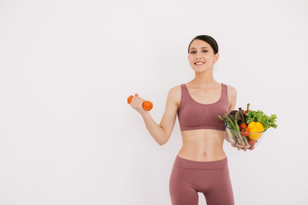 Piękna szczęśliwa młoda kobieta z tacą różnych zdrowych warzyw i pokazująca jej mięśnie