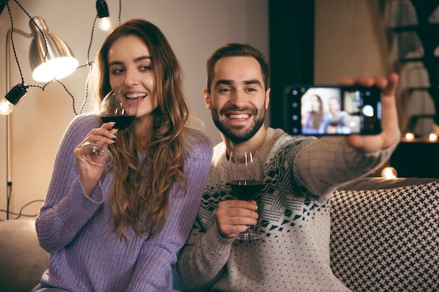 Piękna Szczęśliwa Młoda Para Spędza Razem Romantyczny Wieczór W Domu, Pijąc Czerwone Wino, Biorąc Selfie