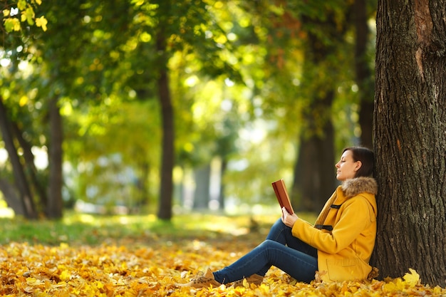 Piękna szczęśliwa uśmiechnięta brązowowłosa kobieta w żółtym płaszczu i dżinsach siedzi pod klonem z czerwoną książką w parku miejskim jesieni w ciepły dzień. Jesienne złote liście. Koncepcja czytania