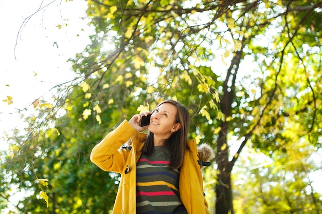 Piękna szczęśliwa wesoła brązowowłosa kobieta w żółtym płaszczu i pasiastym długim rękawie rozmawia przez telefon komórkowy w jesiennym parku miejskim w ciepły dzień. Jesienne złote liście.