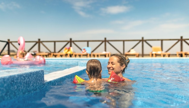 Piękna szczęśliwa wesoła mama bawi się ze swoim małym zabawnym, uroczym synkiem z jasnymi kolorowymi rękawami w ciemnoniebieskim basenie z czystą przezroczystą wodą, latem