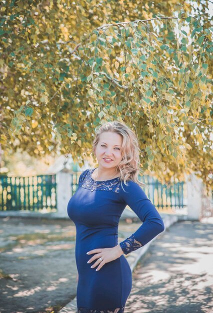piękna szczupła blondynka w długiej niebieskiej sukience uśmiecha się pod jesiennym drzewem