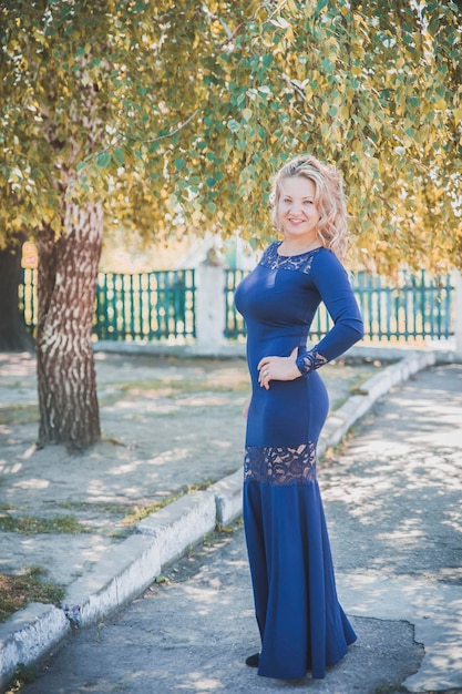piękna szczupła blondynka w długiej niebieskiej sukience uśmiecha się pod jesiennym drzewem