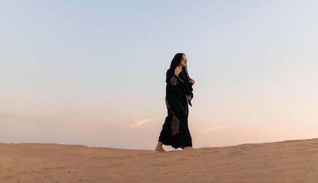 Piękna tajemnicza kobieta w tradycyjnej arabskiej czarnej długiej sukience stoi na pustyni o zachodzie słońca