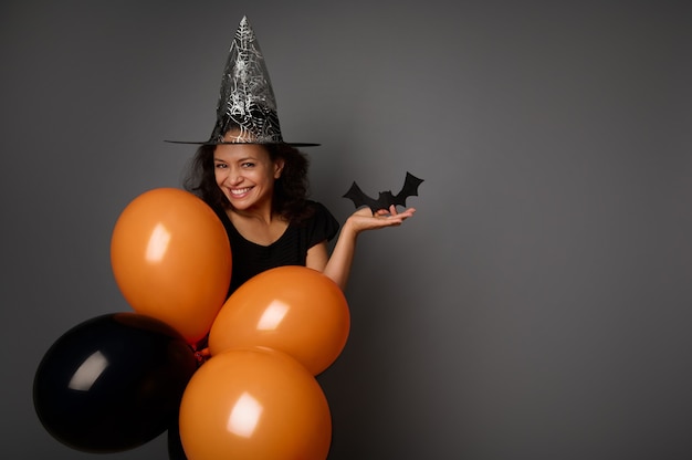 Piękna tajemnicza uśmiechnięta kobieta w kapeluszu czarodzieja, ubrana na czarno, trzyma pomarańczowe czarne balony i ręcznie wycinany z filcu czarny nietoperz, patrzy w kamerę. Koncepcja Halloween party na szarym tle, kopia przestrzeń
