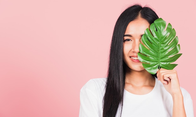 Piękna twarz. Portret azjatyckiej pięknej młodej kobiety ze świeżą zdrową skórą trzyma zielony liść monstera na twarzy, liście tropikalne, studio strzał odizolowane na różowym tle, koncepcja spa do pielęgnacji ciała skóry