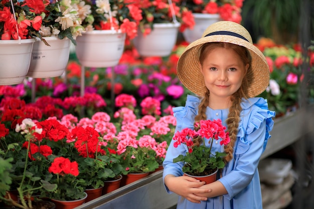 Piękna urocza dziewczyna w słomkowym kapeluszu trzymająca doniczkę z różowymi kwiatami w szklarni z kolorowymi kwiatami