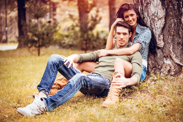 Piękna urocza para siedzi na trawie obok drzewa i cieszy się w słonecznym parku w jesiennych kolorach. Patrząc na aparat.