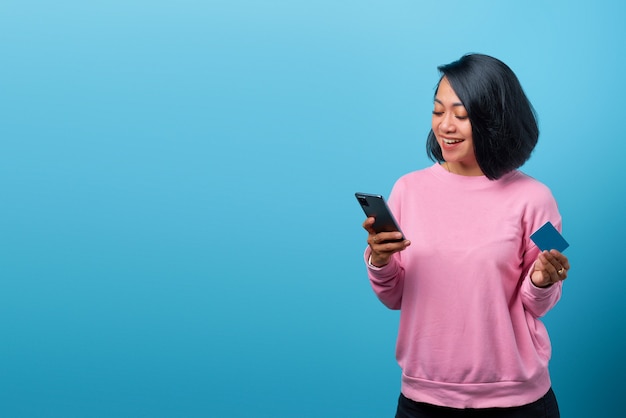 Piękna uśmiechnięta azjatycka kobieta korzystająca z telefonu komórkowego i trzymająca kartę kredytową na niebieskim tle