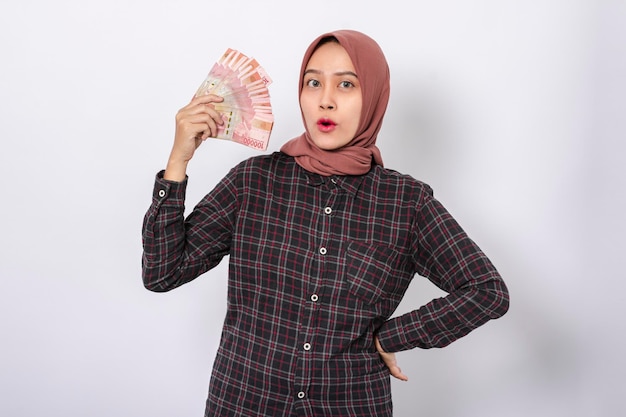 Piękna, uśmiechnięta azjatycka kobieta ubrana w hidżab i flanelową koszulę, trzymającą pieniądze z rupii indonezyjskiej