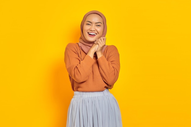 Piękna, uśmiechnięta Azjatycka kobieta w brązowym swetrze i hidżab pocierać ręce i patrząc pewnie na białym tle na żółtym tle. koncepcja religijnego stylu życia ludzi