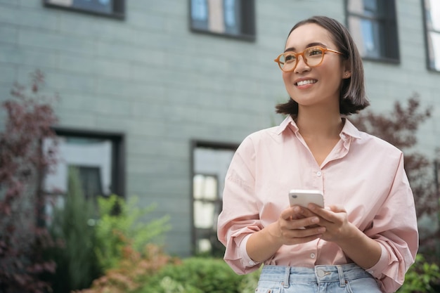 Piękna uśmiechnięta azjatycka kobieta w stylowych okularach trzymająca wiadomości tekstowe z telefonu komórkowego
