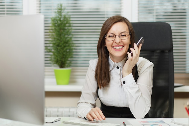 Piękna uśmiechnięta biznesowa kobieta w garniturze i okularach siedząca przy biurku, pracująca przy współczesnym komputerze z dokumentami w jasnym biurze, rozmawiająca przez telefon komórkowy, prowadząca przyjemną rozmowę