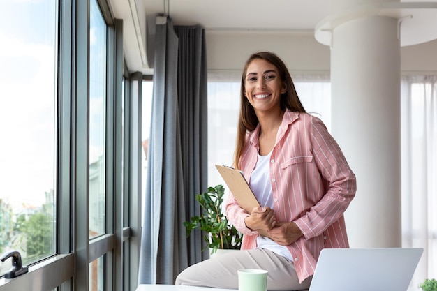 Piękna uśmiechnięta bizneswoman czyta coś na laptopie, opierając się o stół w biurze