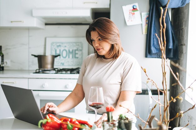 Zdjęcie piękna, uśmiechnięta, brunetka, młoda kobieta z pozytywnym rozmiarem ciała używająca laptopa w kuchni w domu.