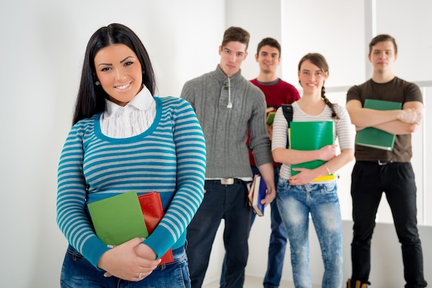 Piękna uśmiechnięta dziewczyna studentka z książką stojącą w sali szkolnej i patrząc na kamery. Za nim szczęśliwa grupa jego przyjaciół.