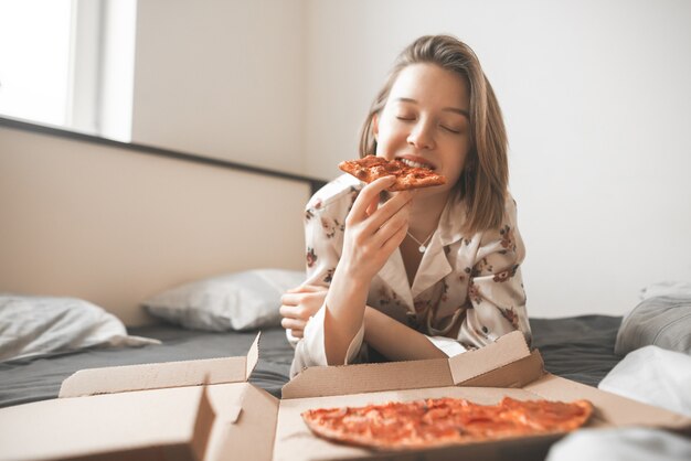 Piękna uśmiechnięta dziewczyna w piżamie leży na łóżku w domu z pudełkiem pizzy