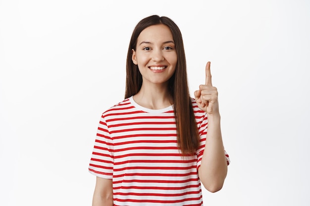 Piękna uśmiechnięta dziewczyna wskazując palcem w górę i patrząc na szczęśliwą, pokazując sprzedaż, tekst promocyjny dla Twojej firmy, stojąc w koszulce na białym tle. Skopiuj miejsce