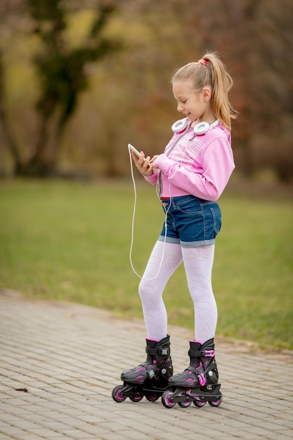 Piękna, Uśmiechnięta Dziewczynka Na Rolkach Po Parku I Przy Użyciu Smartfona.