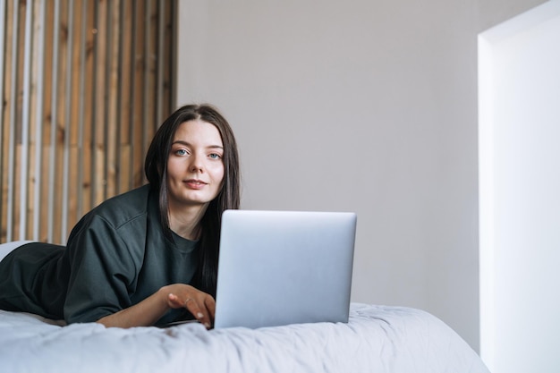 Piękna uśmiechnięta kobieta nastolatek dziewczyna student freelancer z ciemnymi długimi włosami na co dzień przy użyciu laptopa siedzącego na łóżku w domu