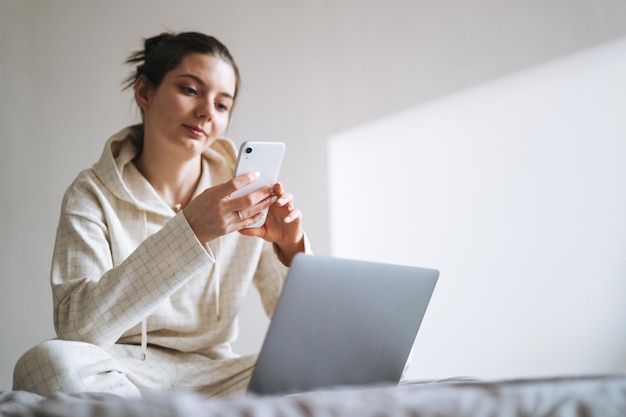 Piękna uśmiechnięta kobieta nastolatek dziewczyna student freelancer z ciemnymi długimi włosami w dorywczo przy użyciu telefonu komórkowego, praca na laptopie, siedząc na kanapie w domu