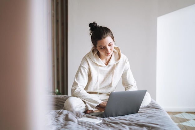 Piękna uśmiechnięta kobieta nastolatek dziewczyna student freelancer z ciemnymi długimi włosami w swobodnym korzystaniu z laptopa siedząc na łóżku w domu