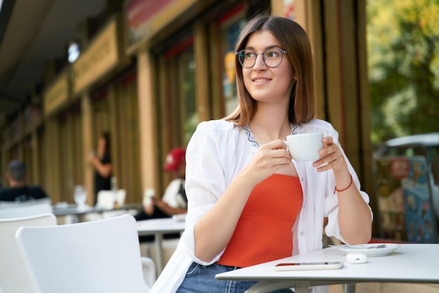 Piękna uśmiechnięta kobieta nosi okulary, trzymając filiżankę kawy w kawiarni. Koncepcja przerwy na kawę