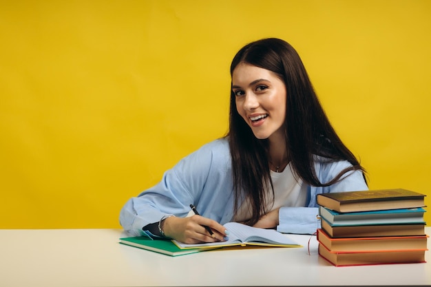 Piękna uśmiechnięta kobieta siedzi przy stole i pisze w swoim notatniku na żółtym tle Koncepcja edukacji