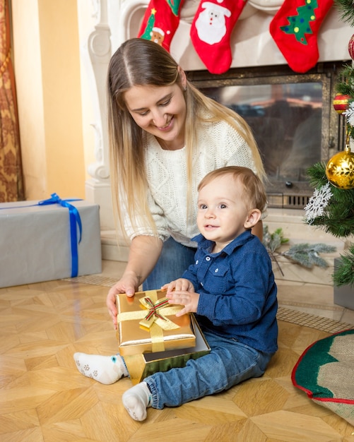 Piękna uśmiechnięta matka i jej synek z prezentem świątecznym na podłodze w salonie