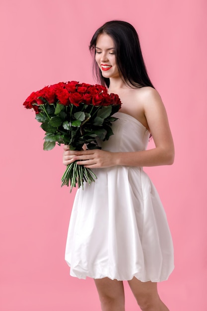 Piękna uśmiechnięta młoda kobieta w białej sukni z dużym bukietem czerwonych róż na różowym tle