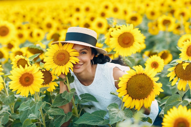 Piękna uśmiechnięta młoda kobieta w kapeluszu z kwiatem na oku i twarzy na polu słoneczników
