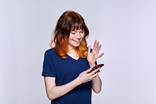 Zdjęcie piękna uśmiechnięta nastolatka patrząca na ekran smartfona na jasnym tle studia pozytywna rudowłosa dziewczyna z telefonem w rękach styl życia technologia komunikacji młodzież młodzi ludzie
