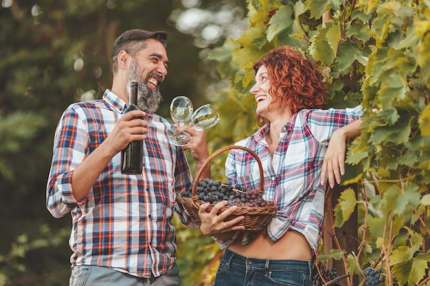 Piękna Uśmiechnięta Para Bawi Się I Degustuje Wino W Winnicy. Mężczyzna Trzyma Butelkę Wina, A Kobieta Kosz Z Winogronami.