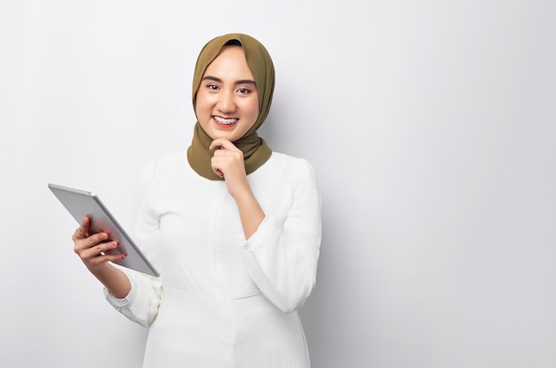 Piękna uśmiechnięta przyjazna młoda arabska azjatycka muzułmanka ubrana w zielony hidżab trzymająca cyfrową tabletkę i kładąca rękę na podbródku odizolowana na białym tle Koncepcja religijnego stylu życia ludzi