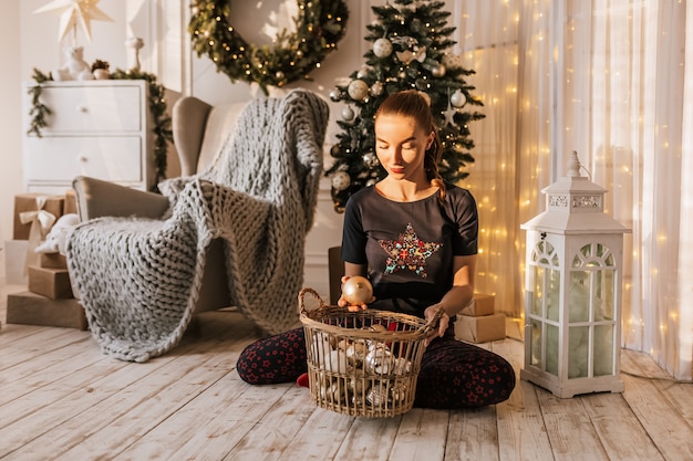Piękna wesoła szczęśliwa młoda dziewczyna na podłodze w piżamie z zabawkami bożonarodzeniowymi w pobliżu drzewa nowego roku w domu