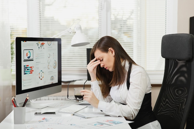 Piękna zmęczona zakłopotana i stresująca brązowe włosy biznesowa kobieta w garniturze i okularach siedząca przy biurku z filiżanką kawy, pracująca przy współczesnym komputerze z dokumentami w jasnym biurze.