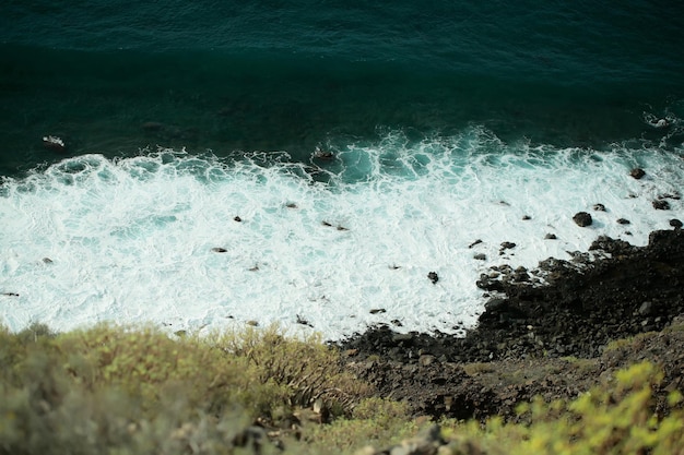 Piękne akwamarynowe fale morskie lub oceaniczne surfują z białą pianką uderzają o czarne skały lub kamienie wzdłuż plaży lub wybrzeża ze szczytu góry w słoneczny dzień na naturalnym tle