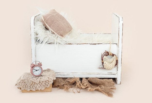 Piękne białe drewniane łóżko do sesji zdjęciowej dla noworodka z futerkiem, poduszką i dekoracją zegara. Małe zaprojektowane miejsce na zdjęcie niemowlęcia na jasnoróżowym tle