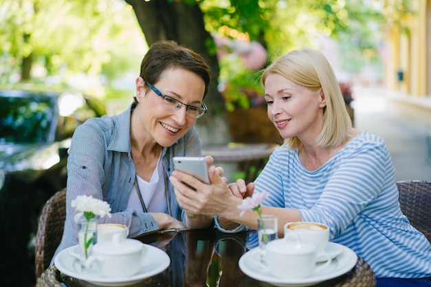 Piękne dojrzałe kobiety za pomocą smartfona rozmawiają śmiejąc się w ulicznej kawiarni