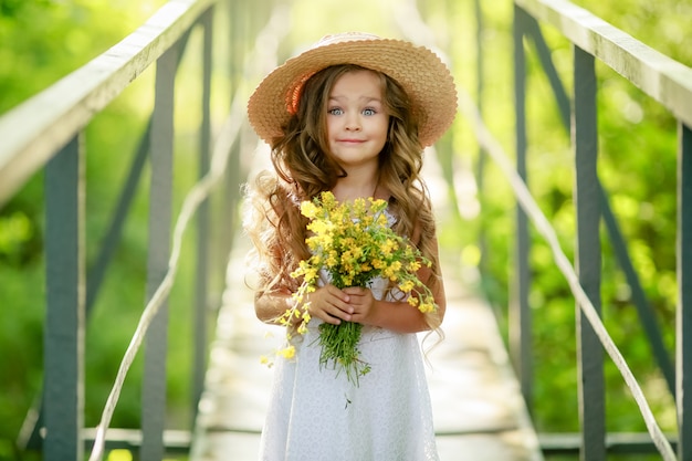 Piękne dziecko dziewczynka ze słomkowym koronkowym kapeluszem na głowie idzie z bukietem żółtych kwiatów leśnym szlakiem