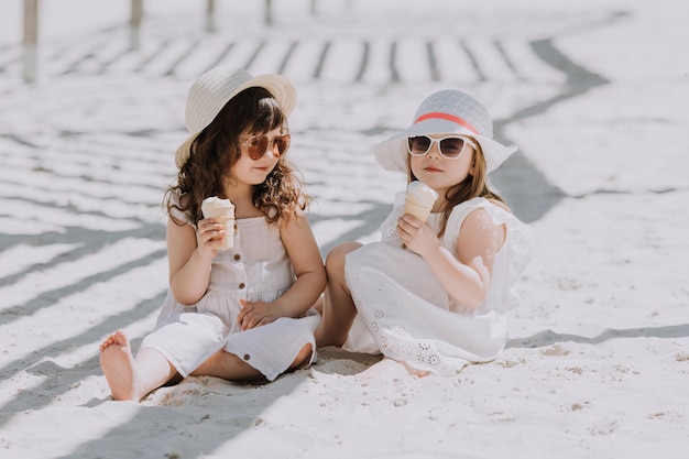 Piękne dziewczynki w białej sukni i kapeluszu jedzą lody na plaży w okresie letnim
