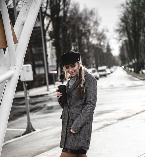 Piękne hipster dziewczyny w stylowy płaszcz, trzymając w ręku filiżankę kawy i odwracając wzrok stojąc na zewnątrz. Młoda modna kobieta pije kawę. Koncepcja miejskiej mody