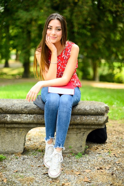 Piękne kobiece college siedzi na ławce w parku i trzymając książkę na kolanach
