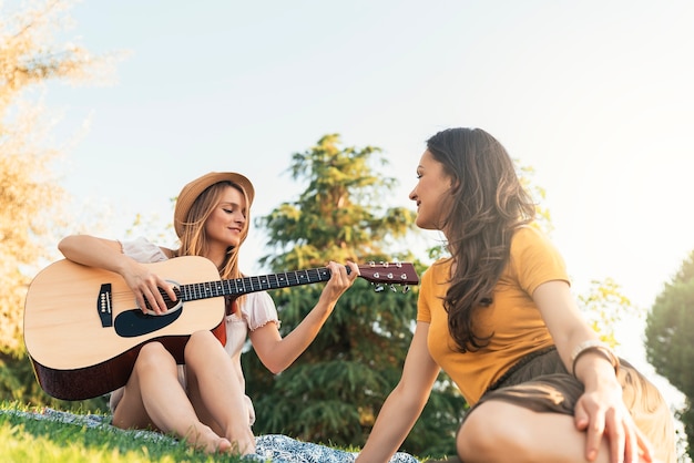 Piękne kobiety zabawy grając na gitarze w parku. Przyjaciele i koncepcja lato.