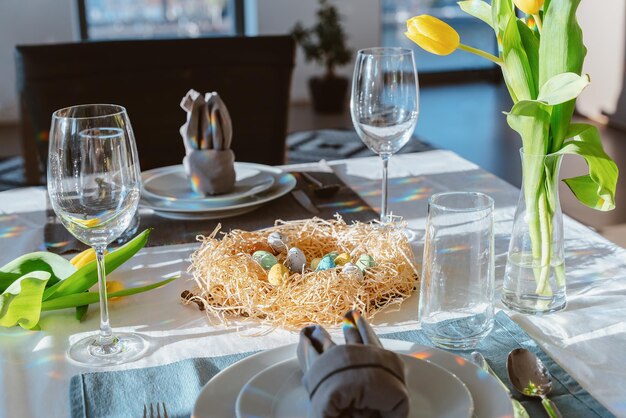 Piękne nakrycie stołu wielkanocnego z świątecznym wystrojem w pomieszczeniu Serwetka w kształcie uszu królika kolorowe jajka w gnieździe świeże żółte tulipany w wazonie Pozdrowienia z pocztówki słonecznej Wnętrze