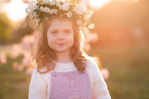 Piękne słodkie dziecko dziewczyna nosi wieniec z kwiatów nad zachodem słońca i kwitnącą wiśnią
