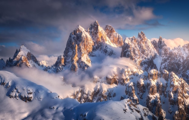 Piękne szczyty górskie w śniegu w zimie o zachodzie słońca Kolorowy krajobraz z wysokimi ośnieżonymi skałami we mgle błękitne niebo z chmurami w zimny wieczór Tre Cime w Dolomitach Włochy Góry alpejskie Natura
