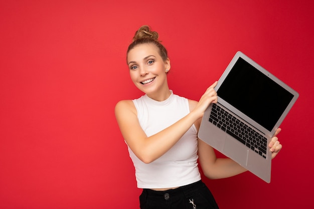 Piękne uśmiechnięte fascynujące szczęśliwe blond młoda kobieta z zebranymi włosami patrząc na kamery posiadania komputera przenośnego na sobie białą koszulkę na białym tle na tle czerwonej ściany.