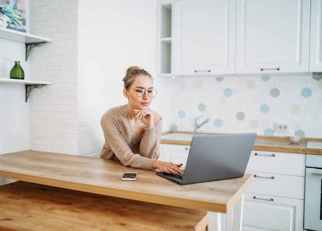 Pięknej uśmiechniętej młodej kobiety uczciwa długie włosy dziewczyna jest ubranym w wygodnym trykotowym pulowerze w szkłach używać laptop przy jaskrawą kuchnią